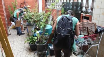 dois agentes estão no jardim de uma casa mexendo em vasos e materiais de ferro empilhados. #paratodosverem
