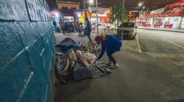 #pracegover Sobre a calçada, mulher entraga cobertor a homem deitado