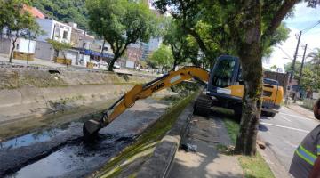 escavadeira parada em calçada recolhe lama de fundo de canal. A escavadeira está na calçada entre duas árvores. #paratodosverem