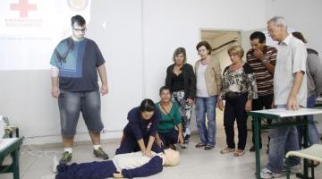 instrutora simula socorro em boneco no chão, pessoas olhando, sala fechada