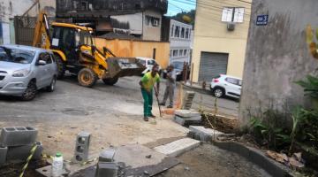 trabalhador realiza obra em rua , escavadeira ao fundo, blocos de concreto #paratodosverem