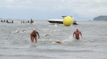 Trecho do mar com nadadores saindo da água, ao fundo uma lancha e uma grande boia amarela #paratodosverem