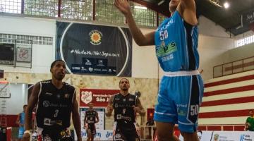 atleta vestido de azul com braço esquerdo esticado junto a uma cesta de basquete