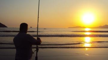 homem segura vara de pesca junto ao mar com o sol ao fundo