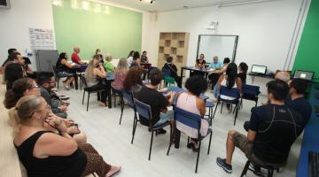 pessoas em uma sala sentadas em cadeiras escolares , ao fundo uma parede verde #paratodosverem