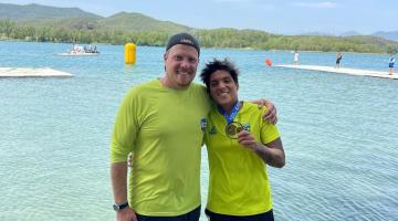 treinador e ana marcela mostrando a medalha com mar ao fundo #paratodosverem