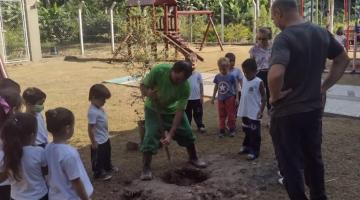 funcionário planta árvore em jardim da escola com várias crianças observando #paratodosverem