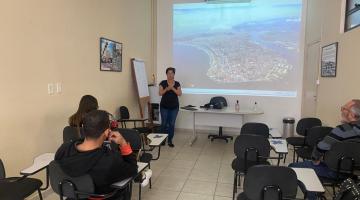 pessoas sentadas em sala de aula observam palestrante com telão ao fundo #paratodosverem