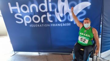 atleta na cadeira de rodas levanta o braço com banner do evento ao fundo #paratodosverem