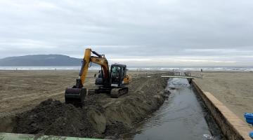 retroescavadeira retira areia ao lado de canal na praia #paratodosverem