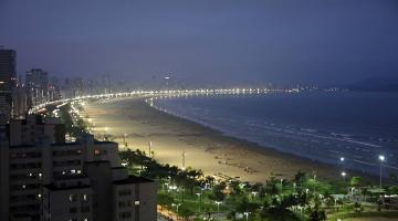 Imagem área da praia a noite, aparecendo prédios, jardins, faixa de areia e mar #paratodosverem