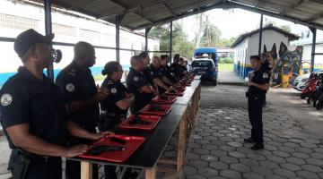 guardas municipais estão em pé atrás de bancada. À frente de todos há bandejas com arma e munição. #paratodosverem