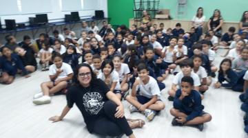 escritora e alunos estão sentados no chão posando para foto. #paratodosverem 