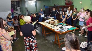 professores estão de pé em uma sala com uma grande mesa com documentos ao centro. #paratodosverem