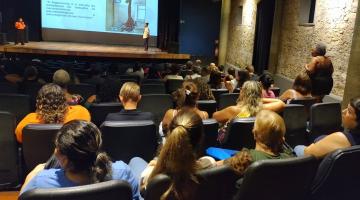 várias pessoas sentadas em auditório assistindo palestra. Ao fundo, no palco, há um telão com imagem. #paratodosverem 