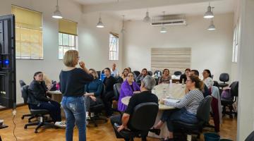 Reunião de várias pessoas que estão sentadas em torno de uma mesa e uma mulher, de costas para a foto, falando ao público. #paratodosverem