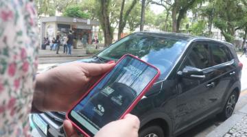 Mulher segura celular com aplicativo do Zona Azul e carro ao fundo. #pratodosverem