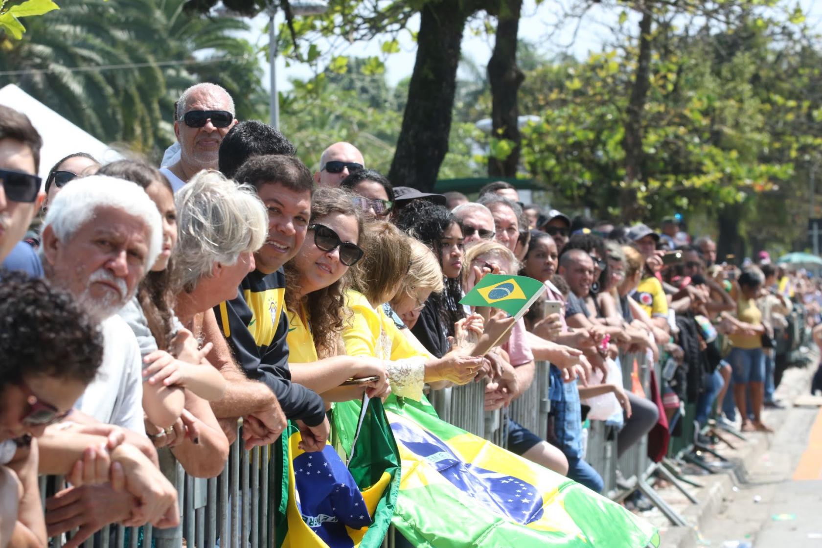 Público está atrás das defesas para assistir ao desfile. Muitas pessoas seguram bandeiras do Brasil. #Pracegover