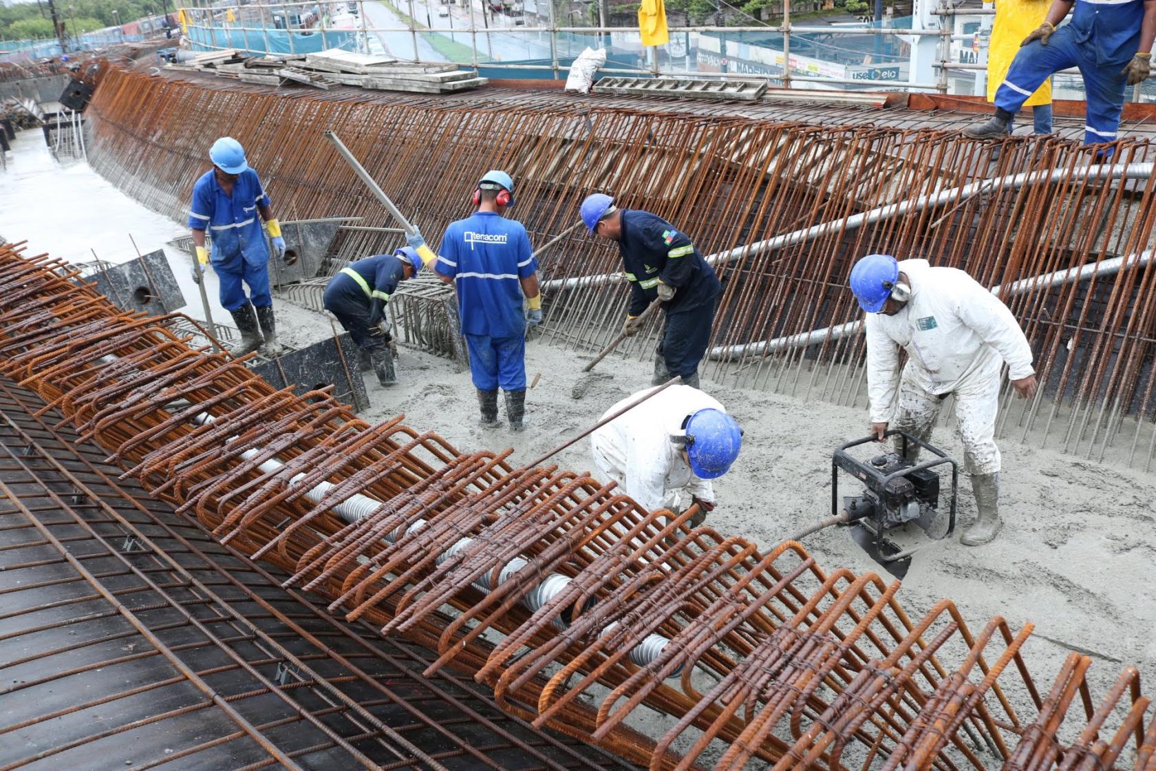 Sete homens trabalham com uniforme e capacete sobre estrutura de concreto e metal do viaduto em construção