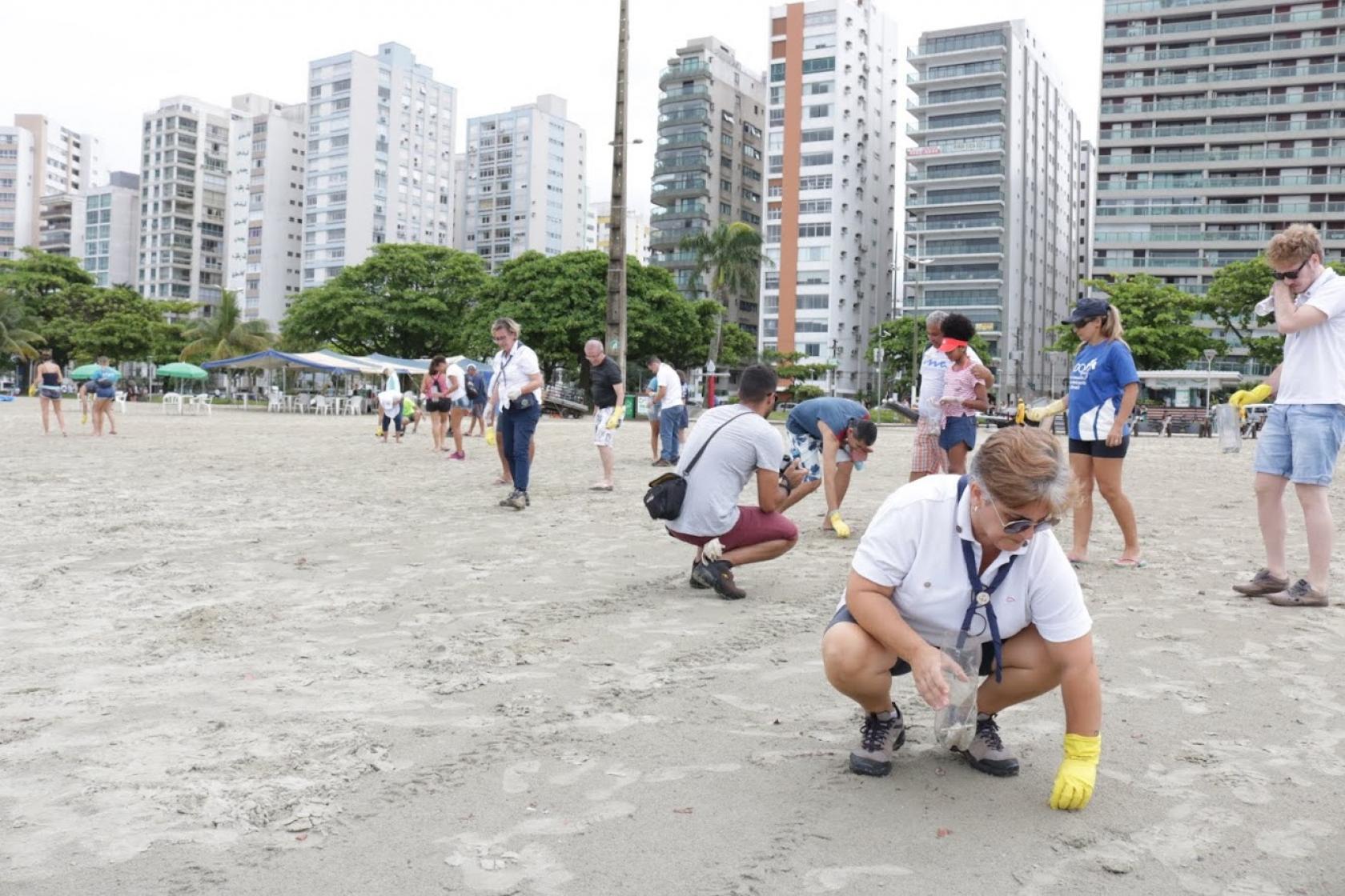 Pessoas caminham e se agacham na areia da praia. Elas fazem movimentos para coletar partículas pequenas de lixo. Ao fundo se vê os prédios da orla. #Pracegover