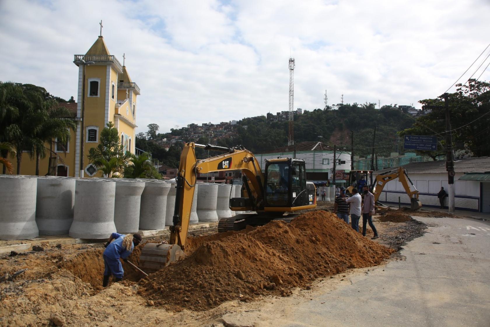 escavadeira e homem atuam em vala com tubos de concreto atrás #paratodosverem 