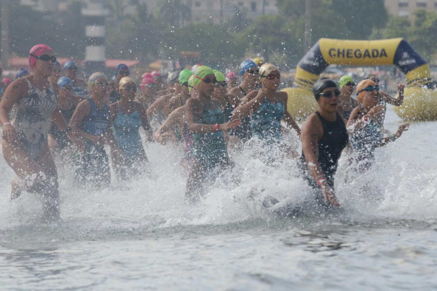 Mulheres entram no mar. A imagem mostra inúmeras mulheres e a água de mexendo com o movimento das atletas. #Paratodosverem