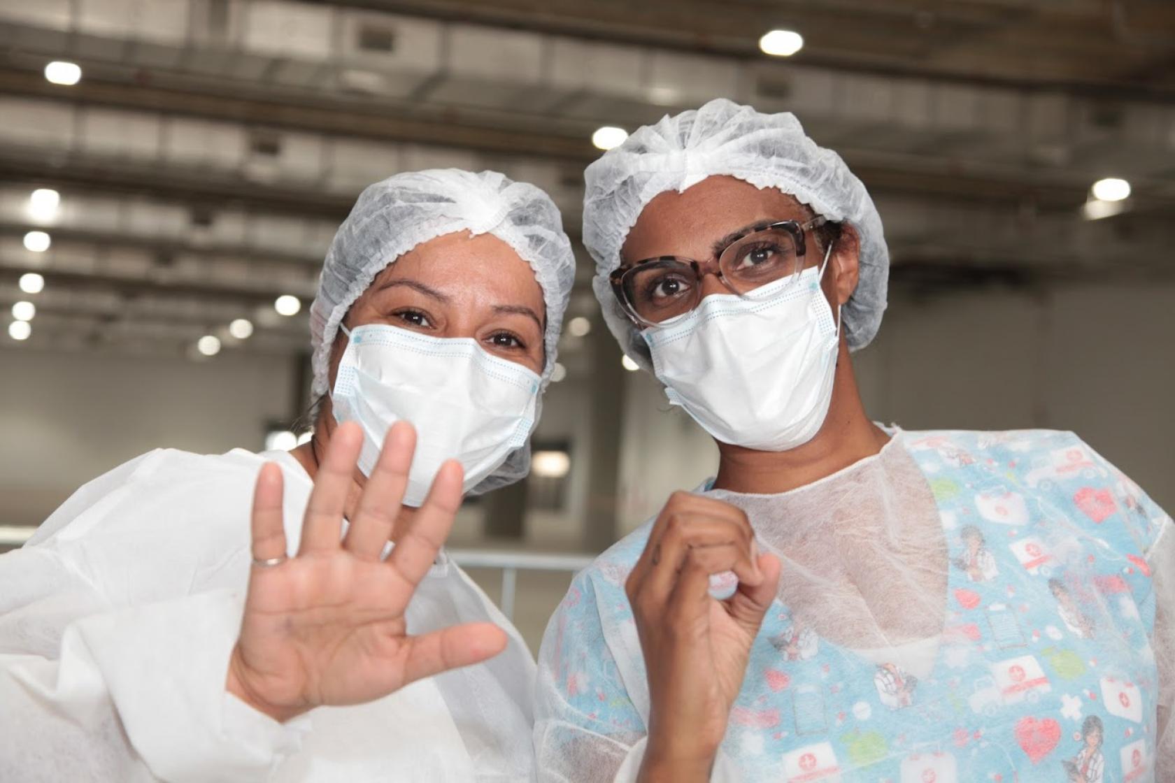 duas mulheres aplicadoras de vacinas posam para foto fazendo o número 50. A da esquerda mostra os cincos dedos da mãos, e a da direita, faz um zero. #paratodosverem