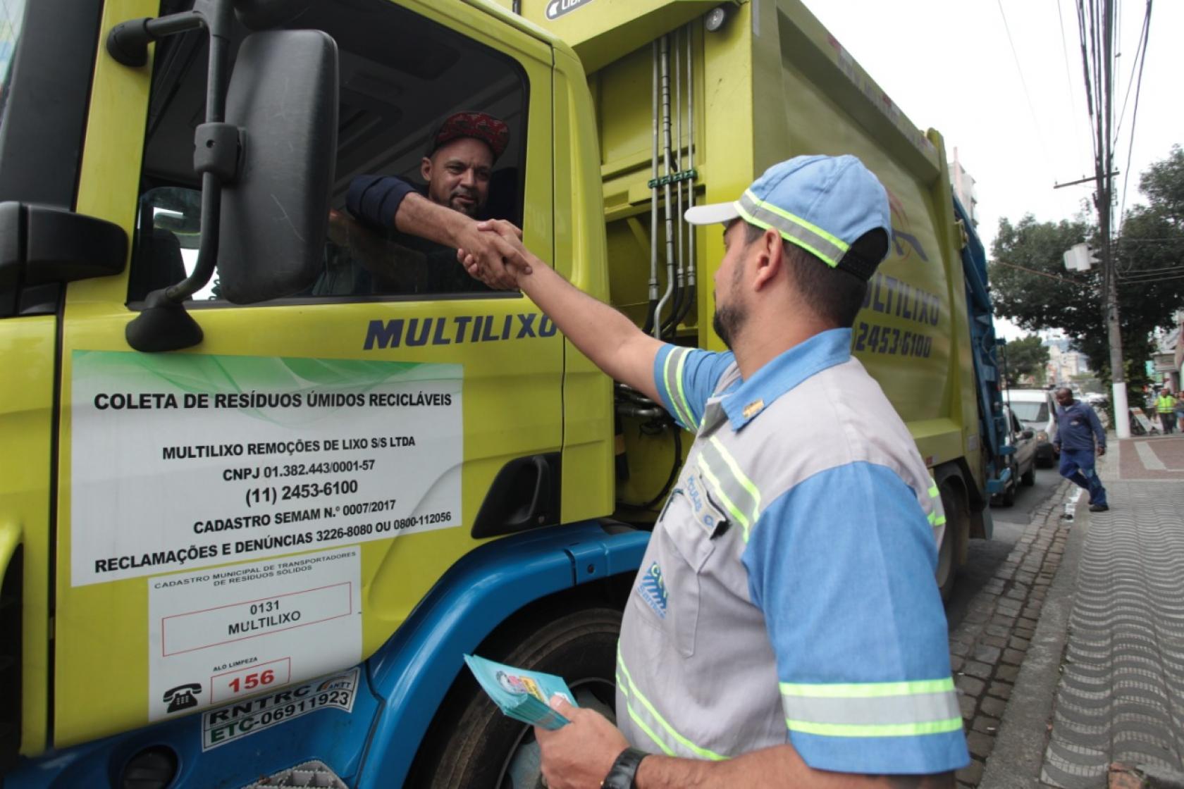 Agente da CET segura folhetos na mão esquerda e cumprimenta motorista de caminhão com a mão direita. O motorista está dentro do veículo. #Pracegover