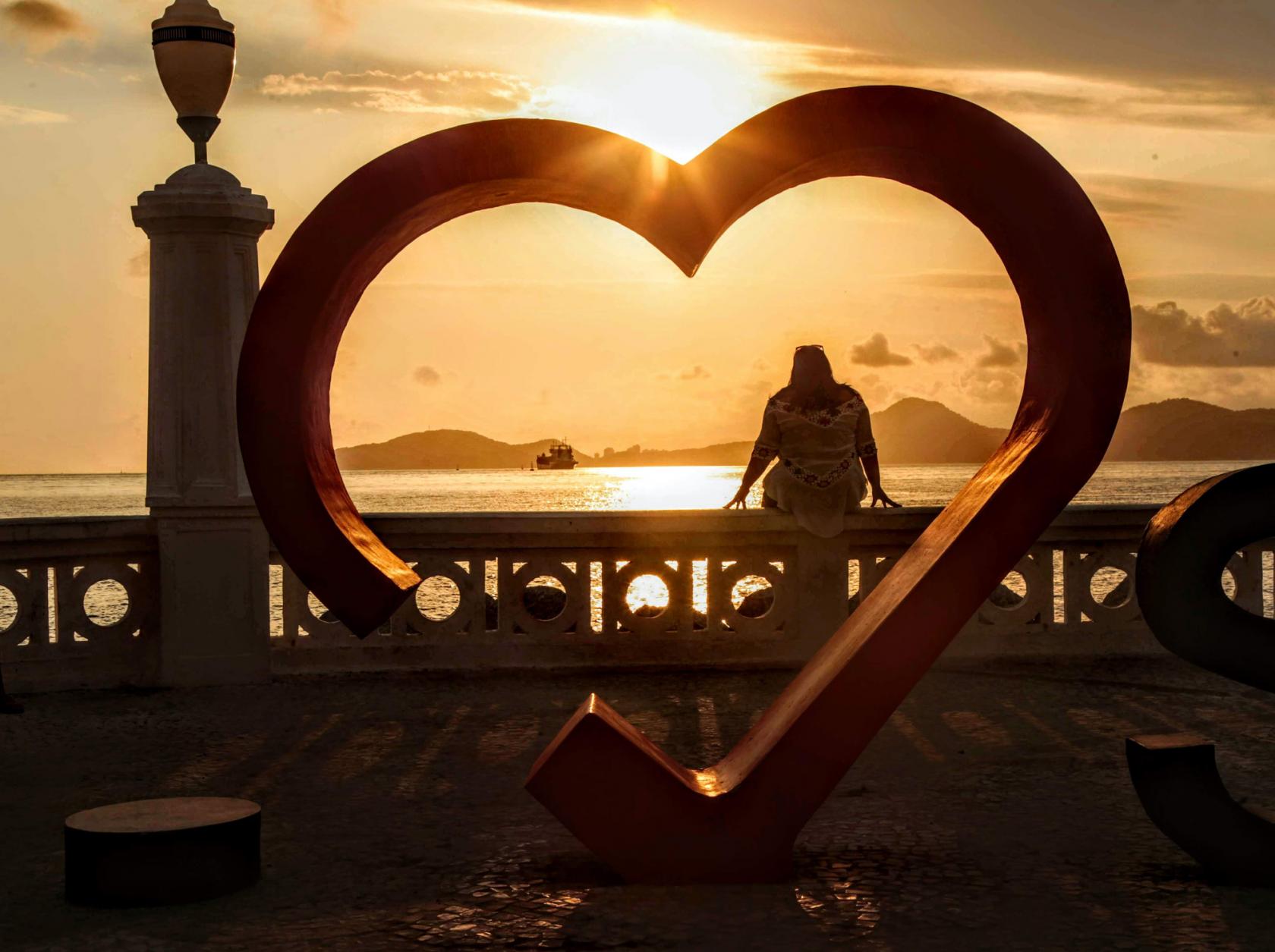 Estrutura em formato de coração na NOva POnta da Praia, em frete a mureta. Há uma pessoa sentada na mureta. O sol está se pondo ao fundo. #Paratodosverem