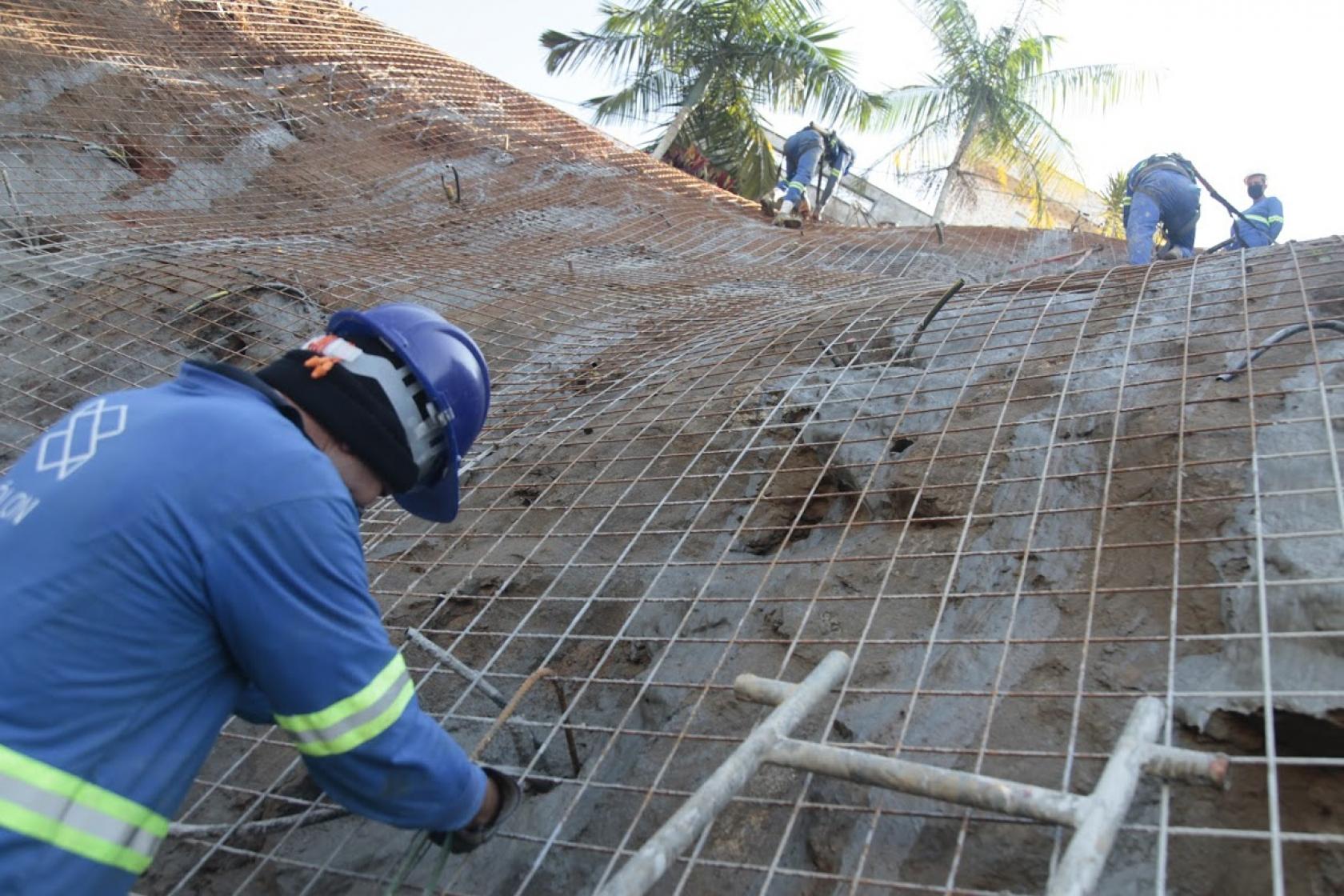 Homens estão trabalhando em encosta de morro. Eles fixam tela de aço na encosta do morro. Um deles está na altura do solo. Outros três estão no alto da estrutura, como se a tivessem escalado. #Paratodosveremécnica do solo grampeado.