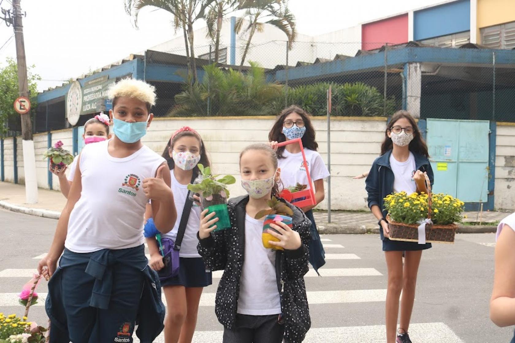 crianças uniformizadas carregam flores pela rua diante de escola. #paratodosverem