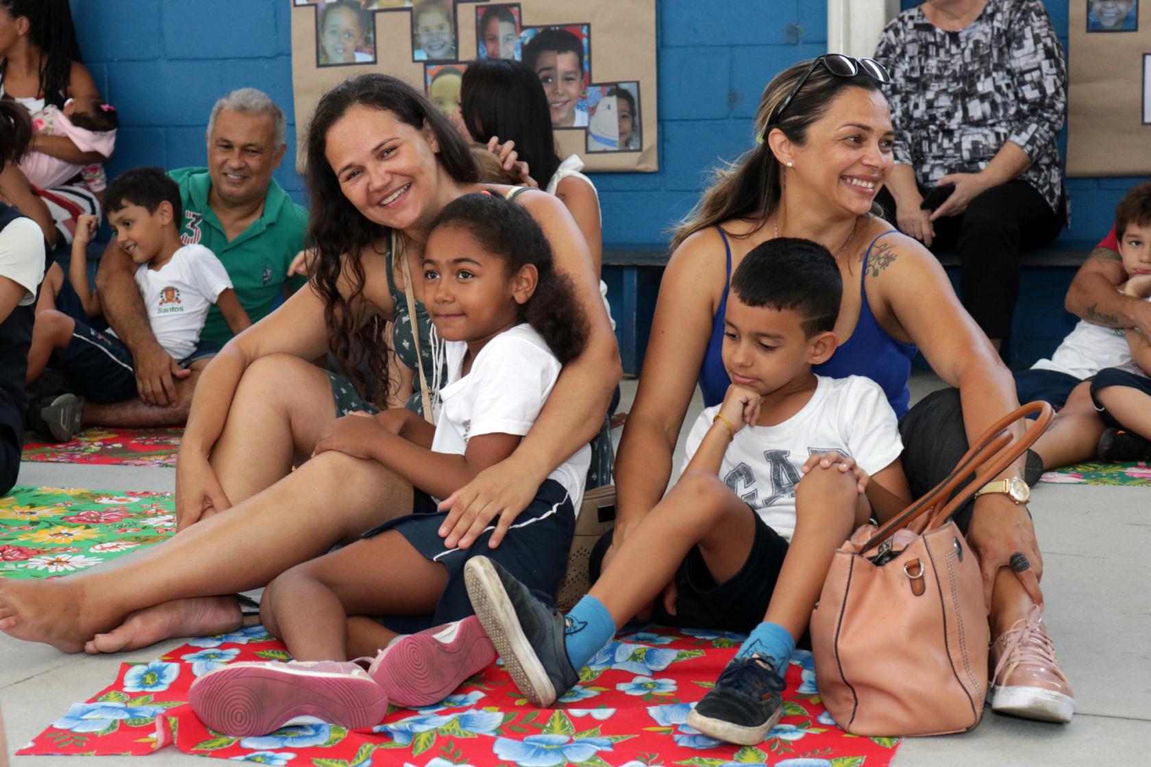 Mães estão sentadas no chão com seus filhos no colo. Todos sorriem. #Pracegover