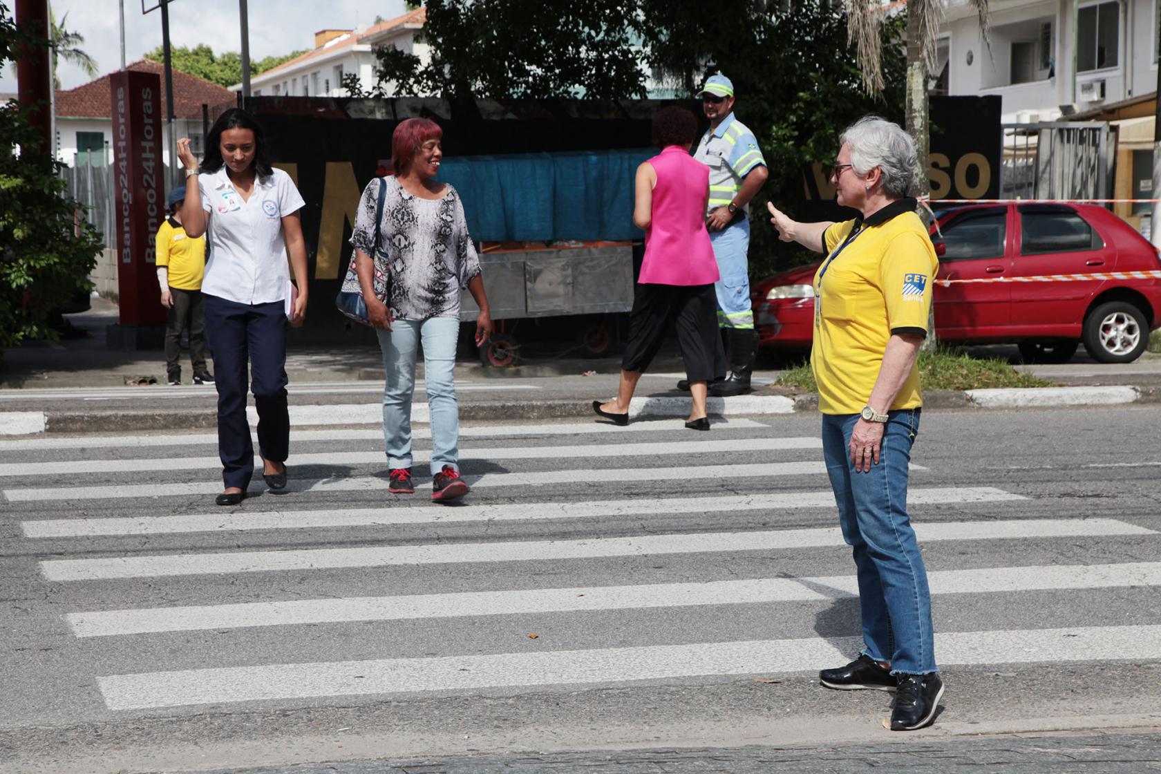 Agente da CET estende a mão mostrando a pessoas como atravessar a rua #paratodosverem