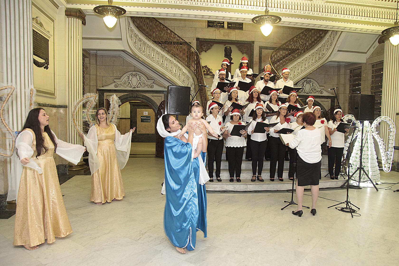 Coral se apresenta na escadaria do hall da prefeitura, regido pela maestrina. Em primeiro plano, uma mulher, representando Maria, segura um bebê, o menino Jesus. Ao lado, anjos acompanham. #Pracegover