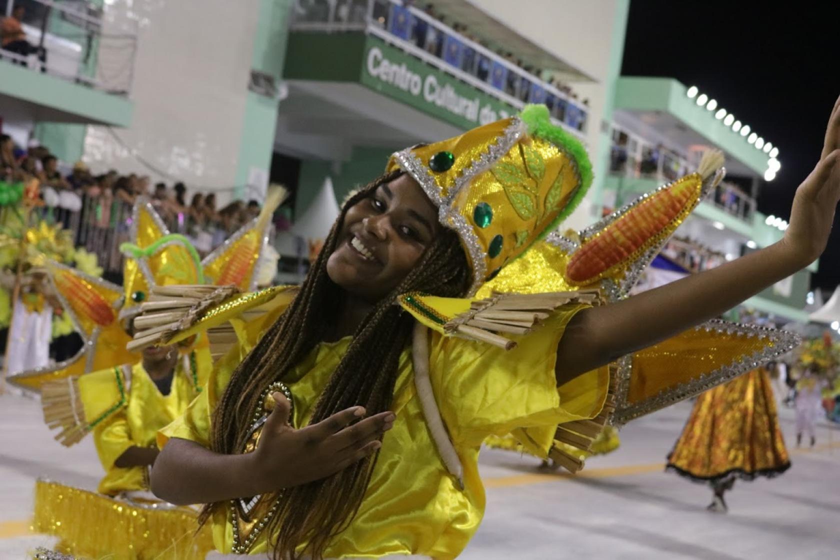 Mulher faz movimento com as mãos para o alto, dançando. Ela tem um adereço na cabeça. #Pracegover