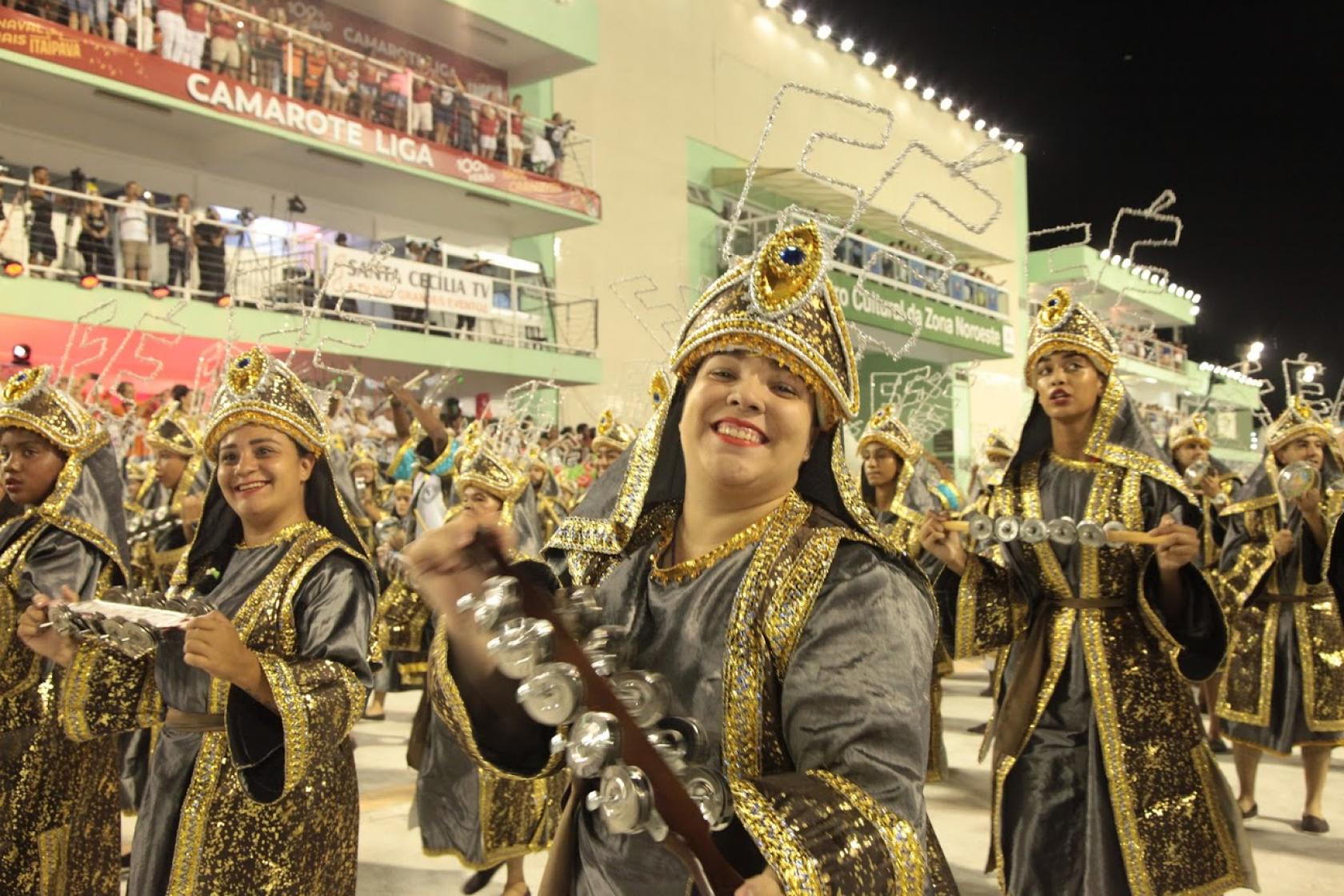 Integrante de bateria movimenta rocar chocalho. Sobre o adereço em sua cabeça se lê: Fé. Ela sorri para a foto Em volta há vários músicos. Ao fundo está a plateia. #Paratodosverem