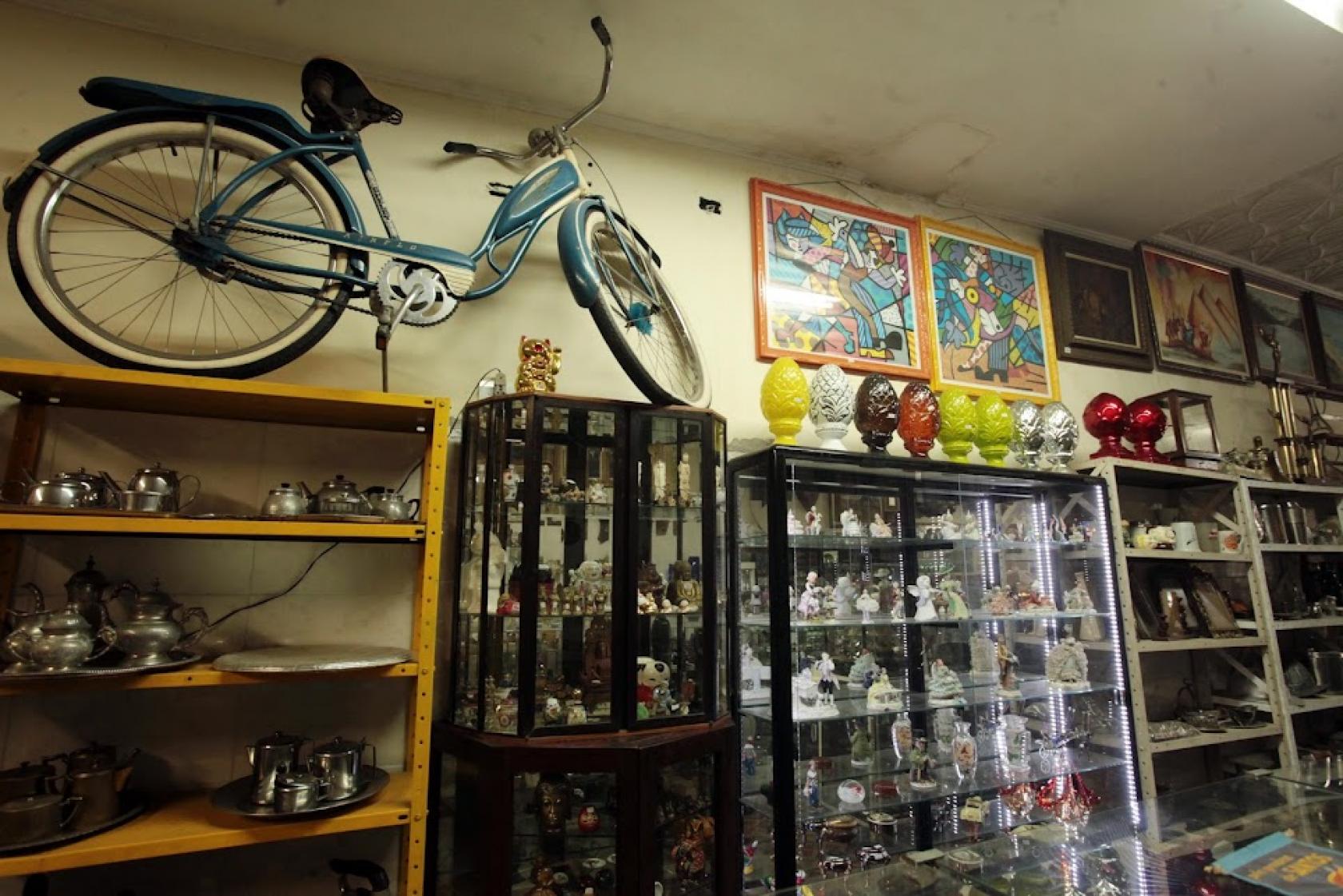 mobiliário antigo em antiquário. Há cristaleiras cheias de objetos, bicicleta e quadros na parede. #paratodosverem