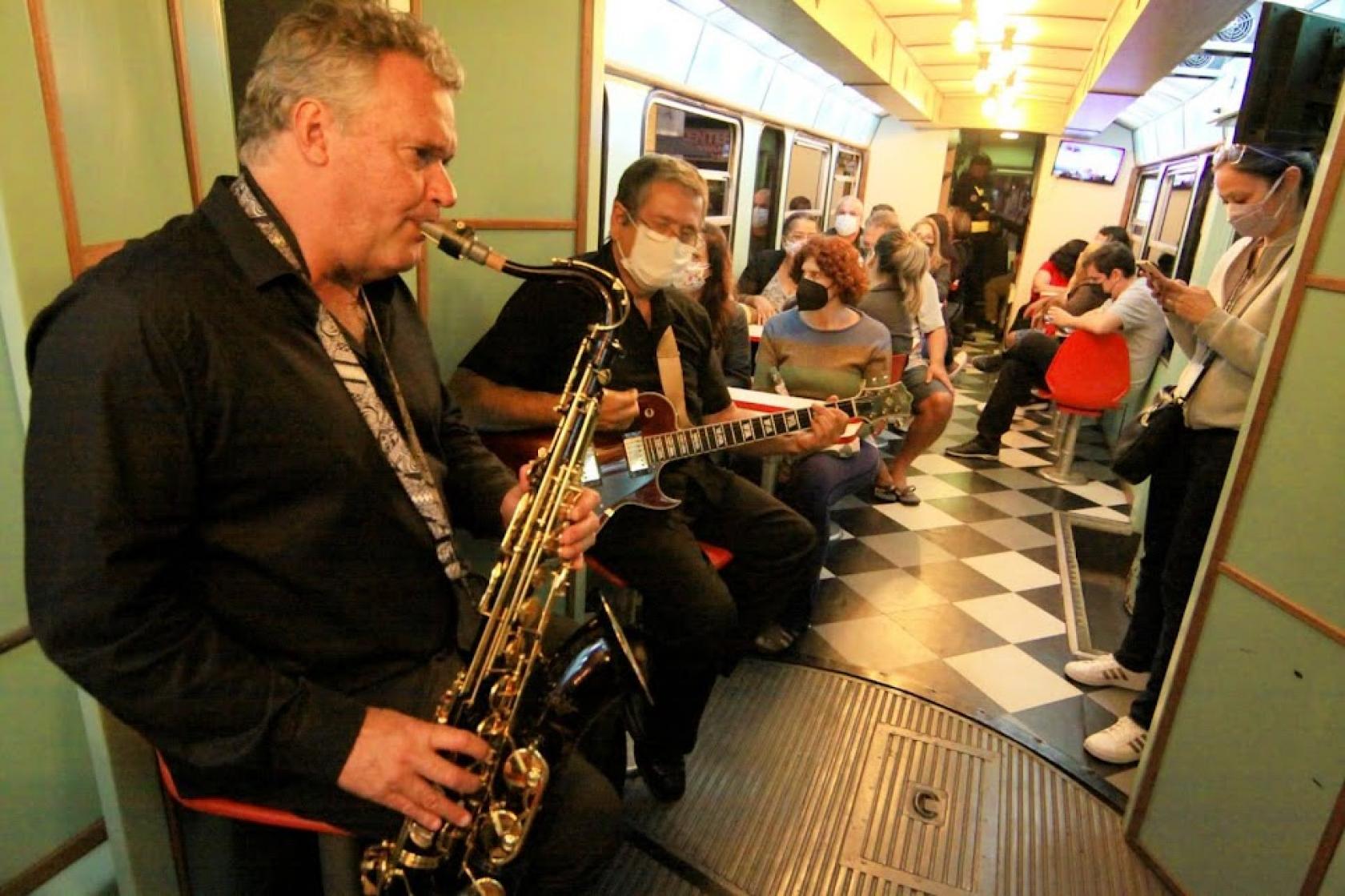 músicos em primeiro plano à esqueda. São dois. Um toca sax e o outro, guitarra. Ao fundo, o público está sentado. #paratodosverem