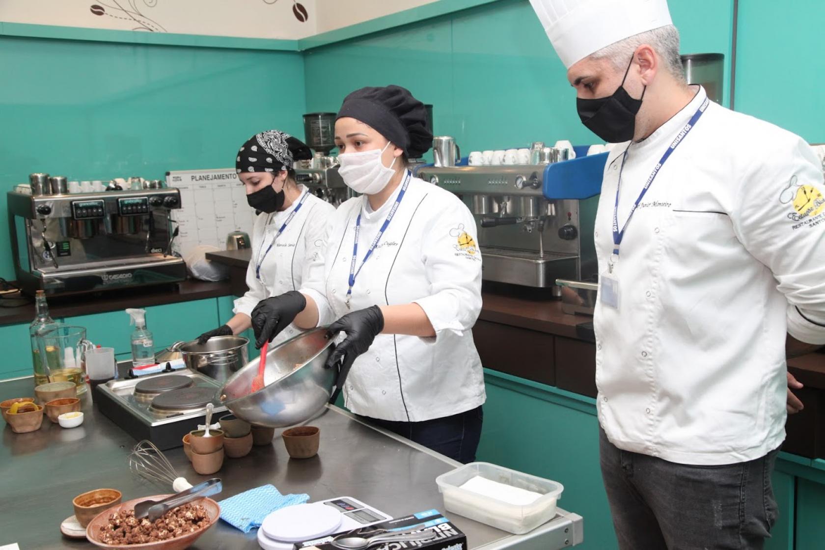 duas mulheres e um homem vestidos de culinaristas estão atrás de mesa. As duas estão preparando um prato. #paratodosverem