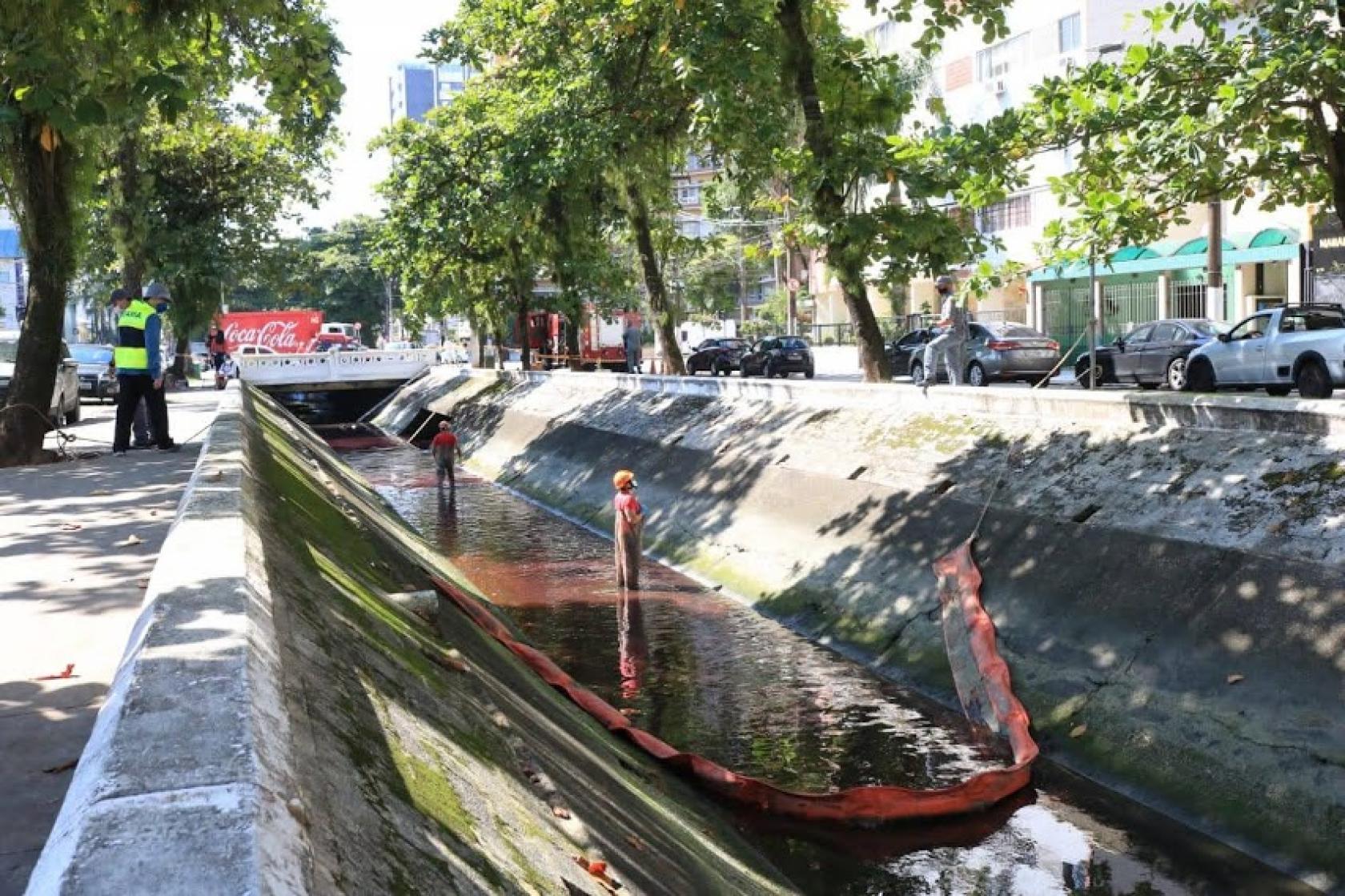 Canal com mancha vermelha. Há uma barreira instalada e dois homens dentro do canal. #paratodosverem