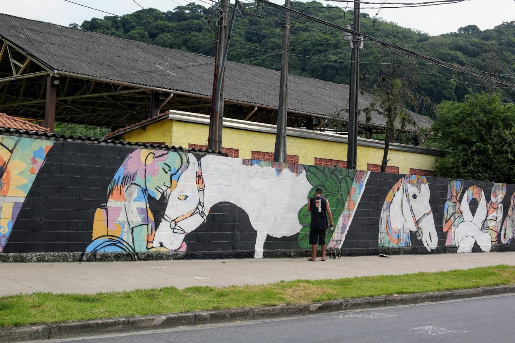 Muro grafitado com desenhos de cavalos. Uma das figuras é a de uma menina em cadeira de rodas com a cabeça apoiada na cabeça do cavalo. #Paratodosverem