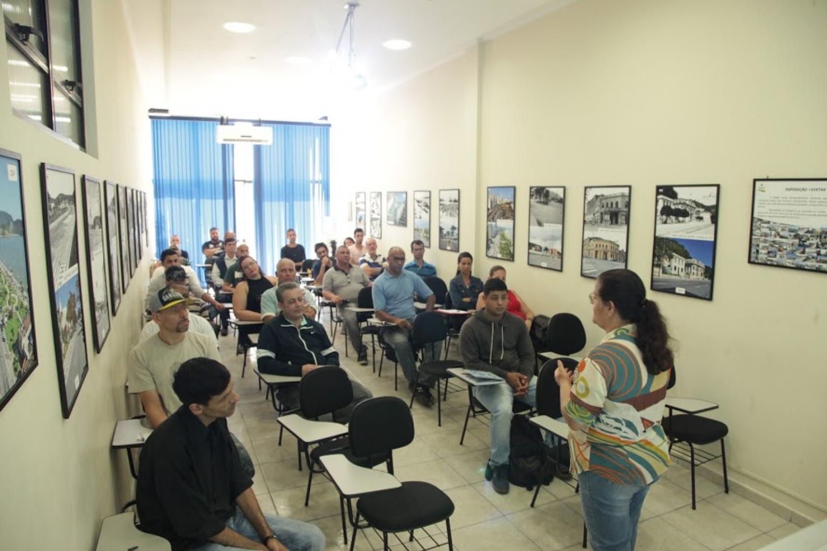 Alunos sentados observam orientadora falando de pé. #pracegover