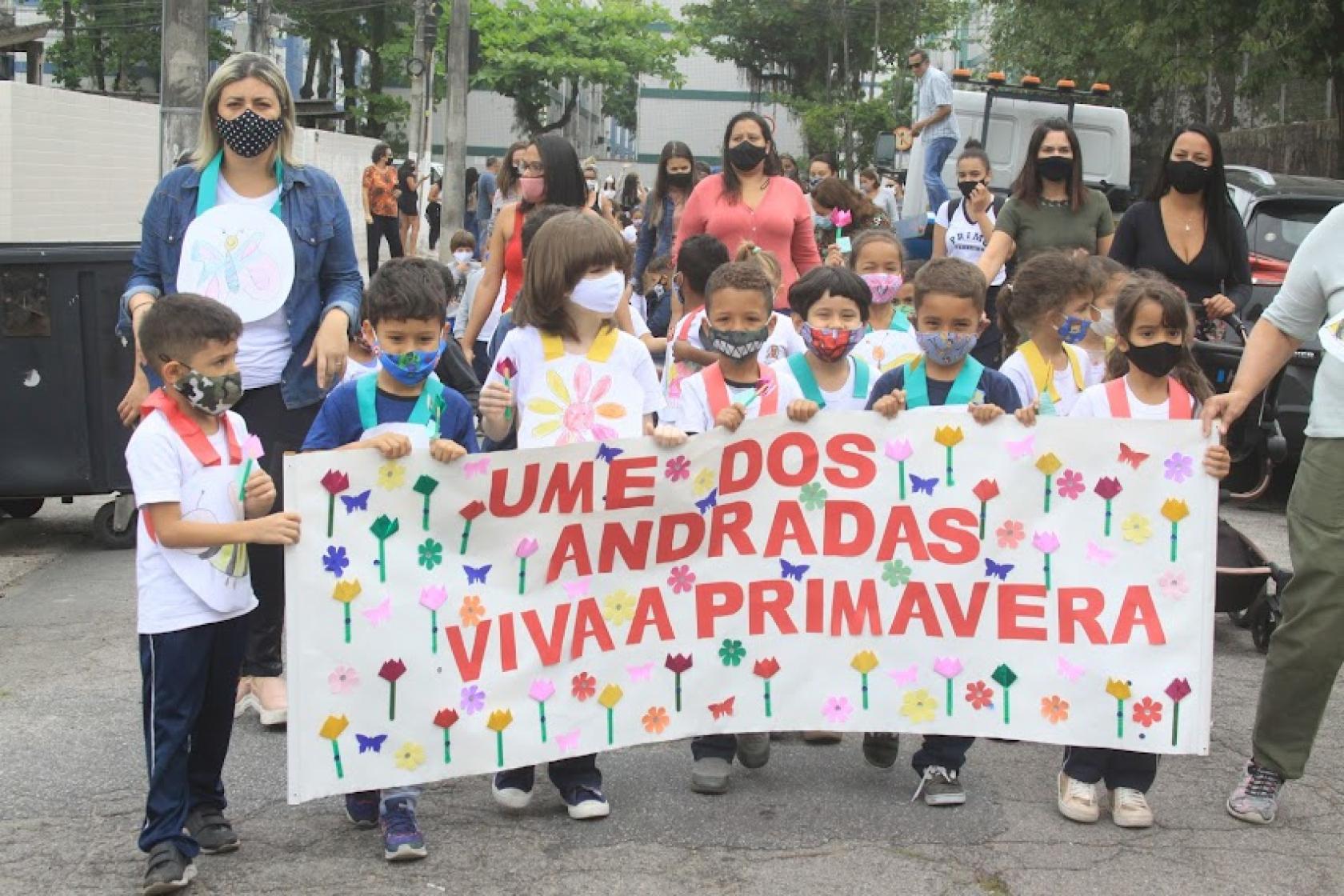 Crianças caminham pela rua, acompanhadas por professoras. Elas carregam uma faixa onde se lê UME DOS ANDRADAS VIVA A PRIMAVERA. #paratodosverem