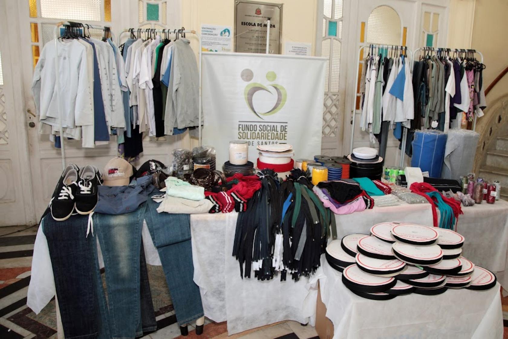 roupas em araras e mesas, tênis e aviamentos. Um banner do fundo social no fundo. #paratodosverem