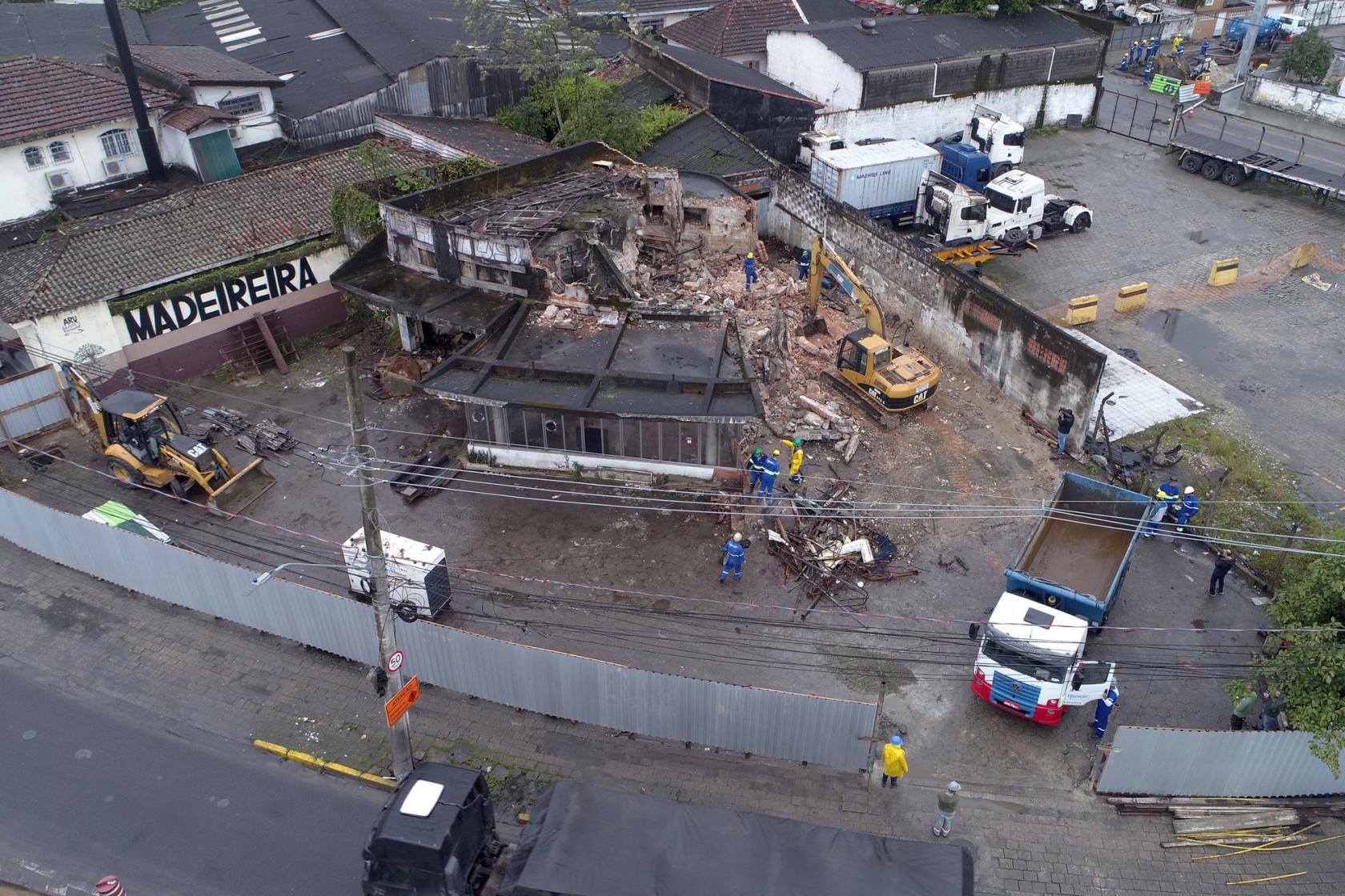 Imagem aérea de antigo posto em demolição. Há máquinas e caminhões na área. Parte da estrutura, ao fundo, já está derrubada. #Pracegover