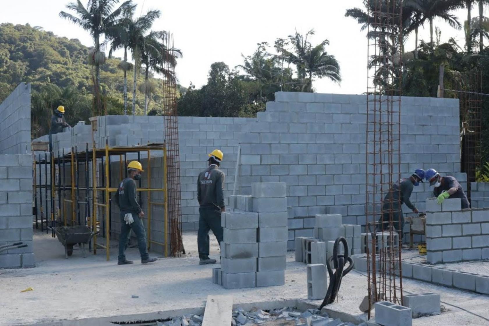 Operários assentam blocos de parede em canteiro de obras. Há estruturas de ferro implantadas no solo. As paredes têm cerca de dois metros de altura. #Paratodosverem