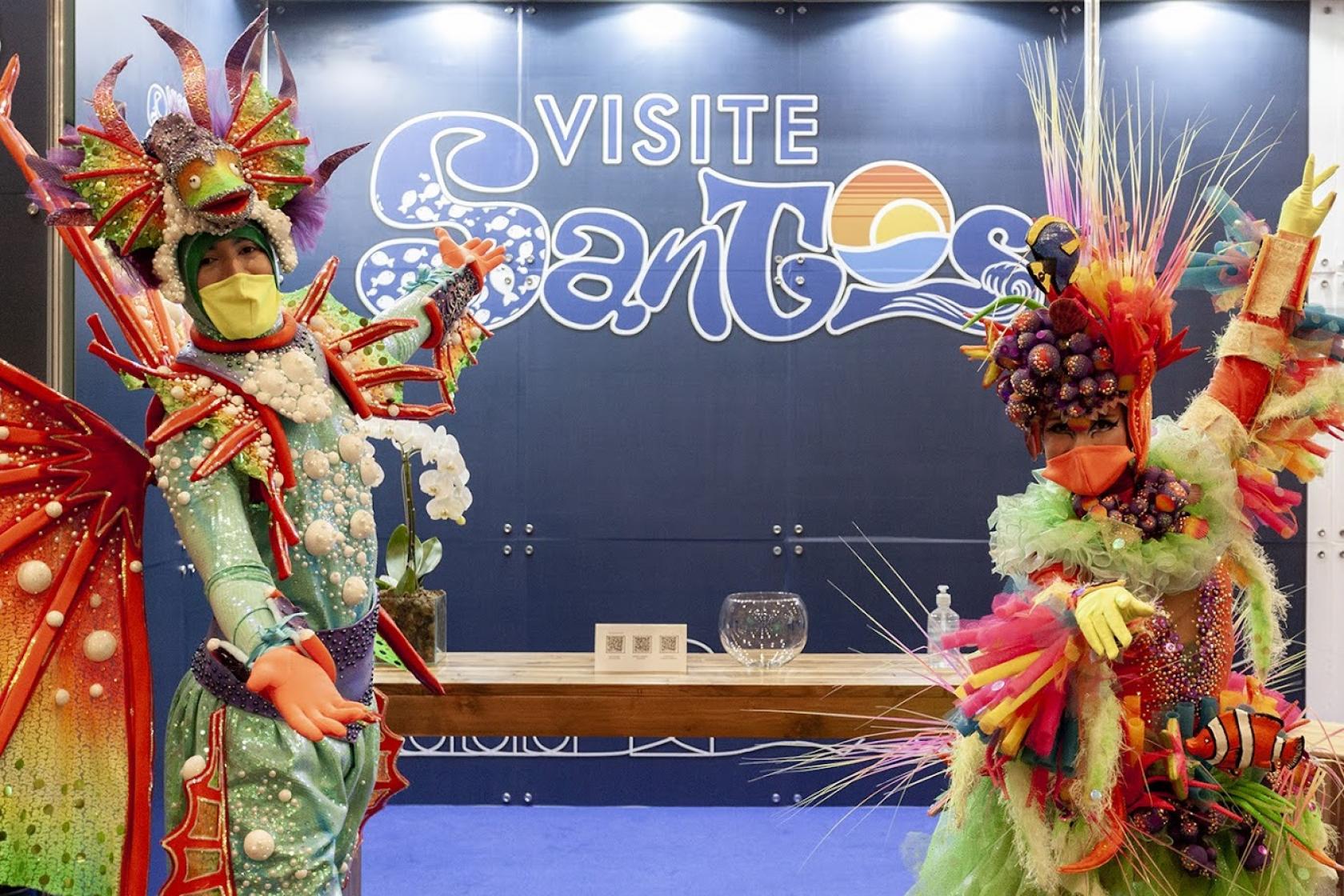 Recepcionistas de estande de Santos vestidos com motivos marinhos coloridos e usando máscaras. #paratodosverem