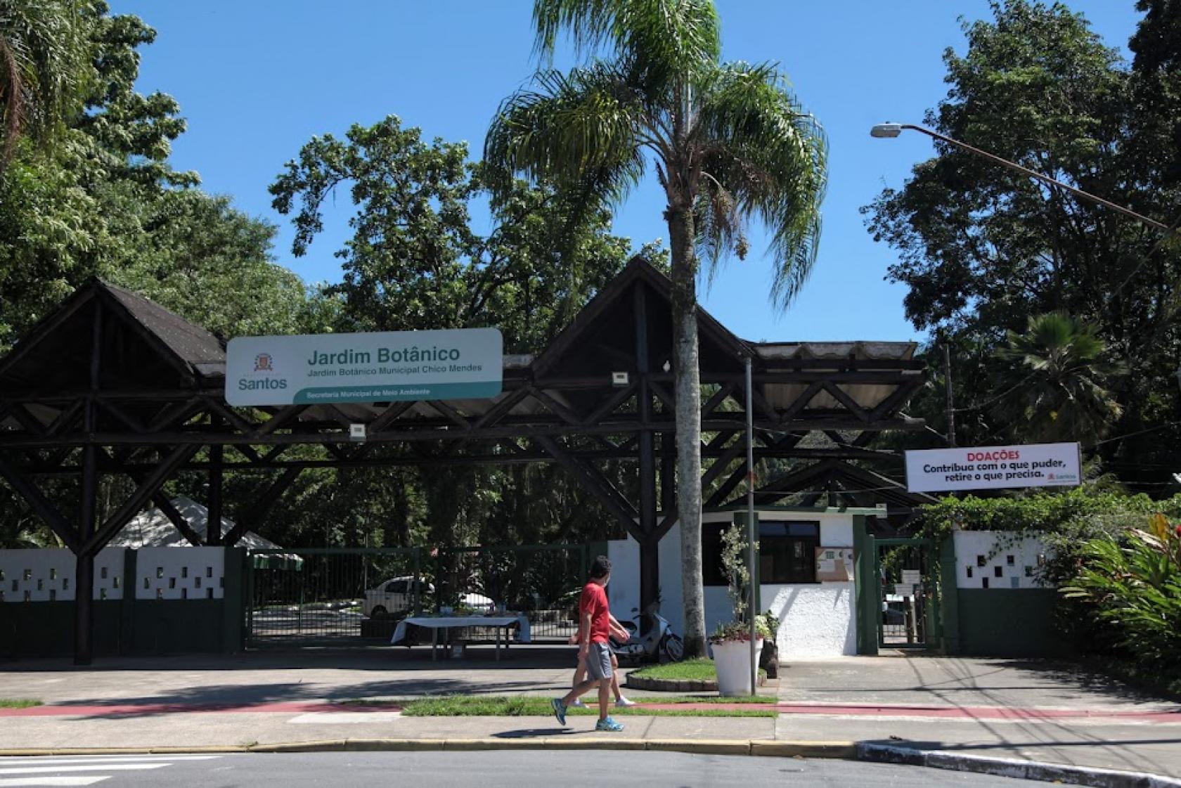 Fachada do jardim botânico com placa, palmeira à frente e árvores variadas ao fundo, atrás do portal de acesso. #paratodosverem