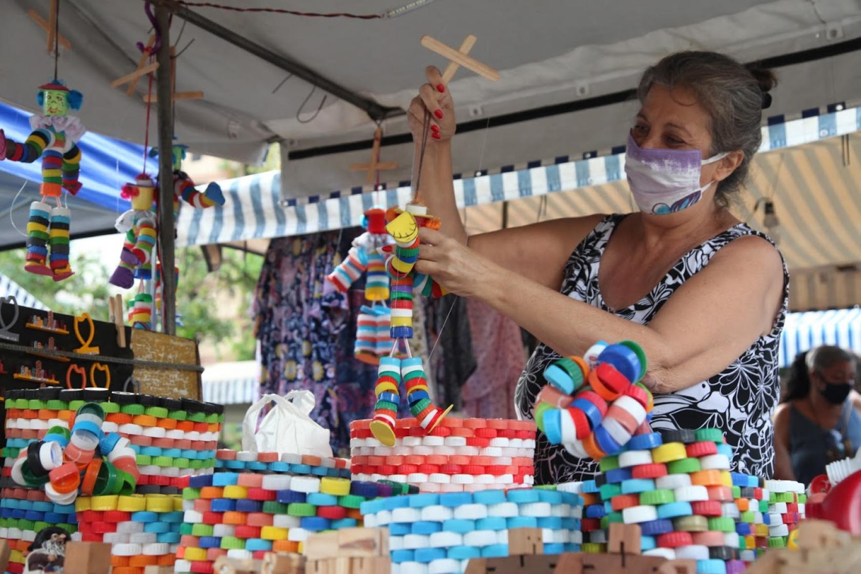 Mulher segura boneco feito de tampas de garrafa. Várias objetos com a mesma peça estão expostos em banca. #Paratodosverem