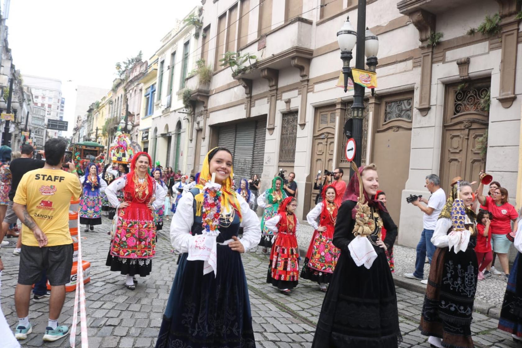 mulheres com roupas t'picas desfilam por rua. #paratodosverem